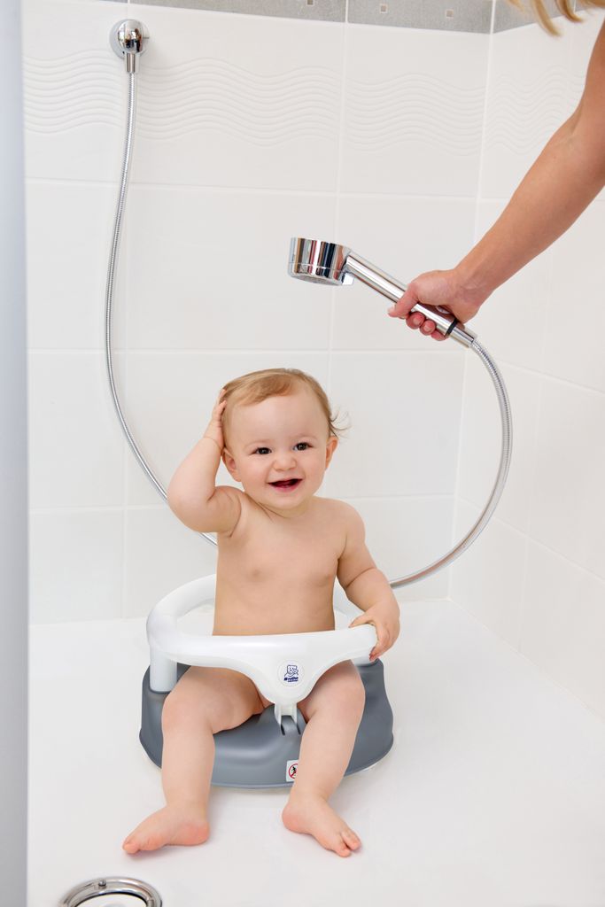 A baby sitting in a bath seat in a shower, with a hand holding a shower head above.