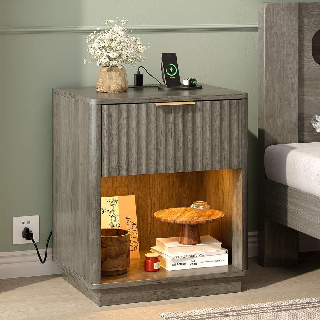 A nightstand with a drawer and an open shelf, placed next to a bed. The nightstand has a charging station on top.