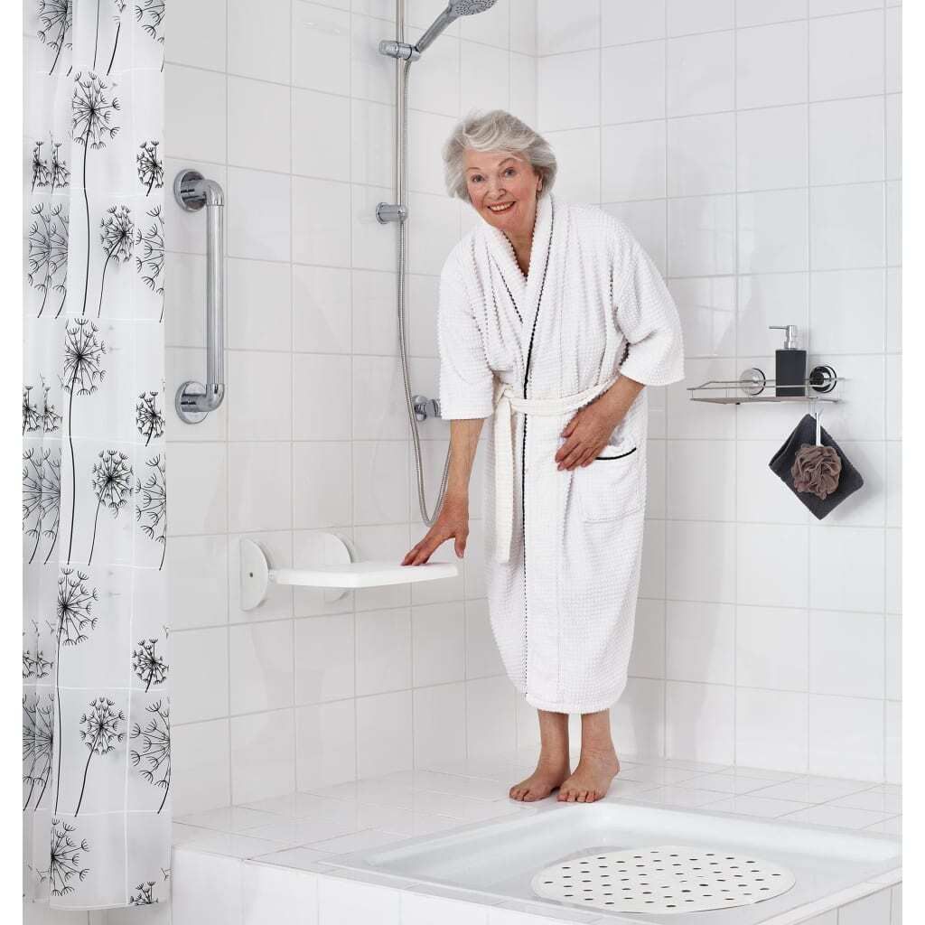 An elderly woman in a white bathrobe stands in a bathroom next to a shower seat.