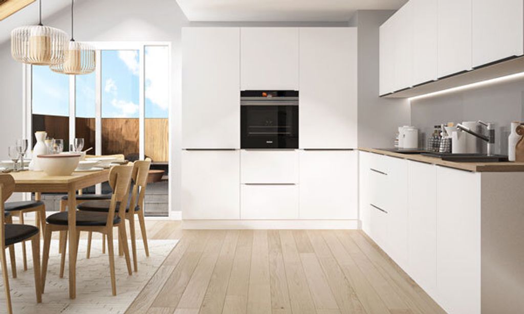 A modern, minimalist kitchen with white cabinets, an integrated oven, and a dining area with a wooden table and chairs.