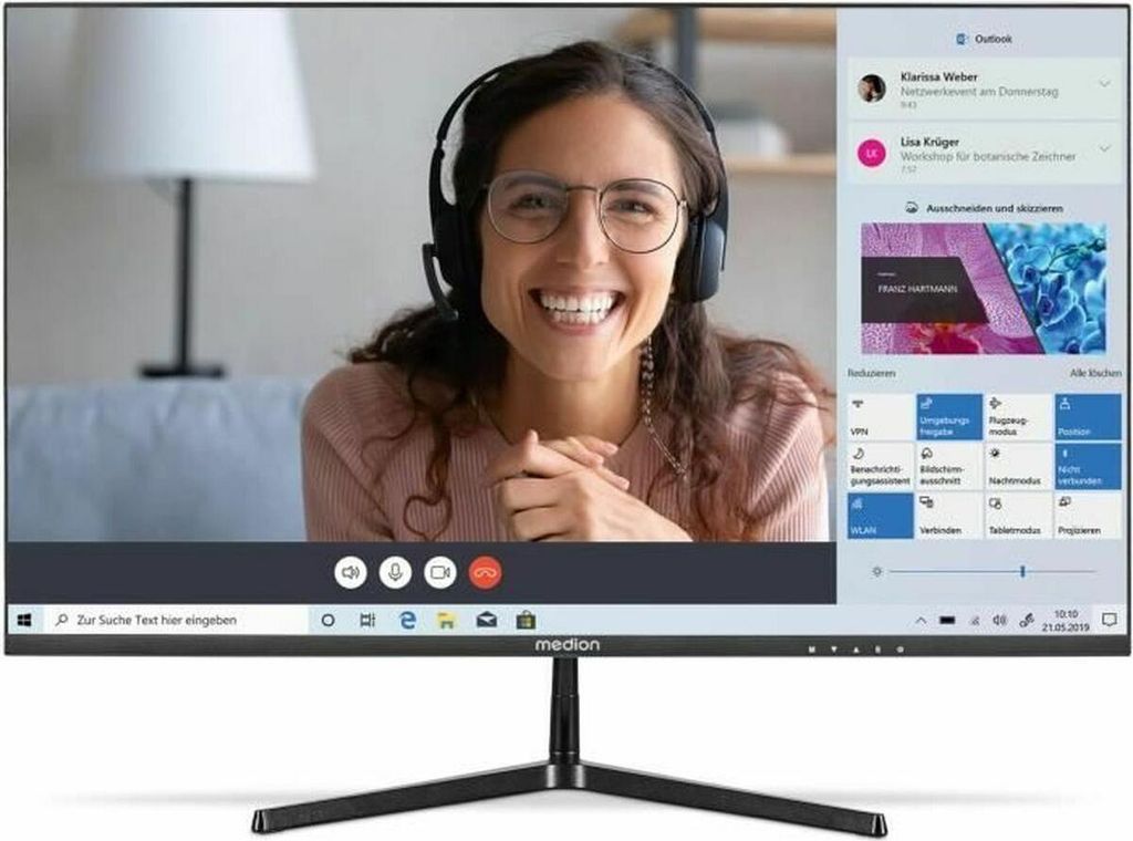 A woman is on a video call on a computer monitor. The monitor screen displays the video call interface and various application windows.