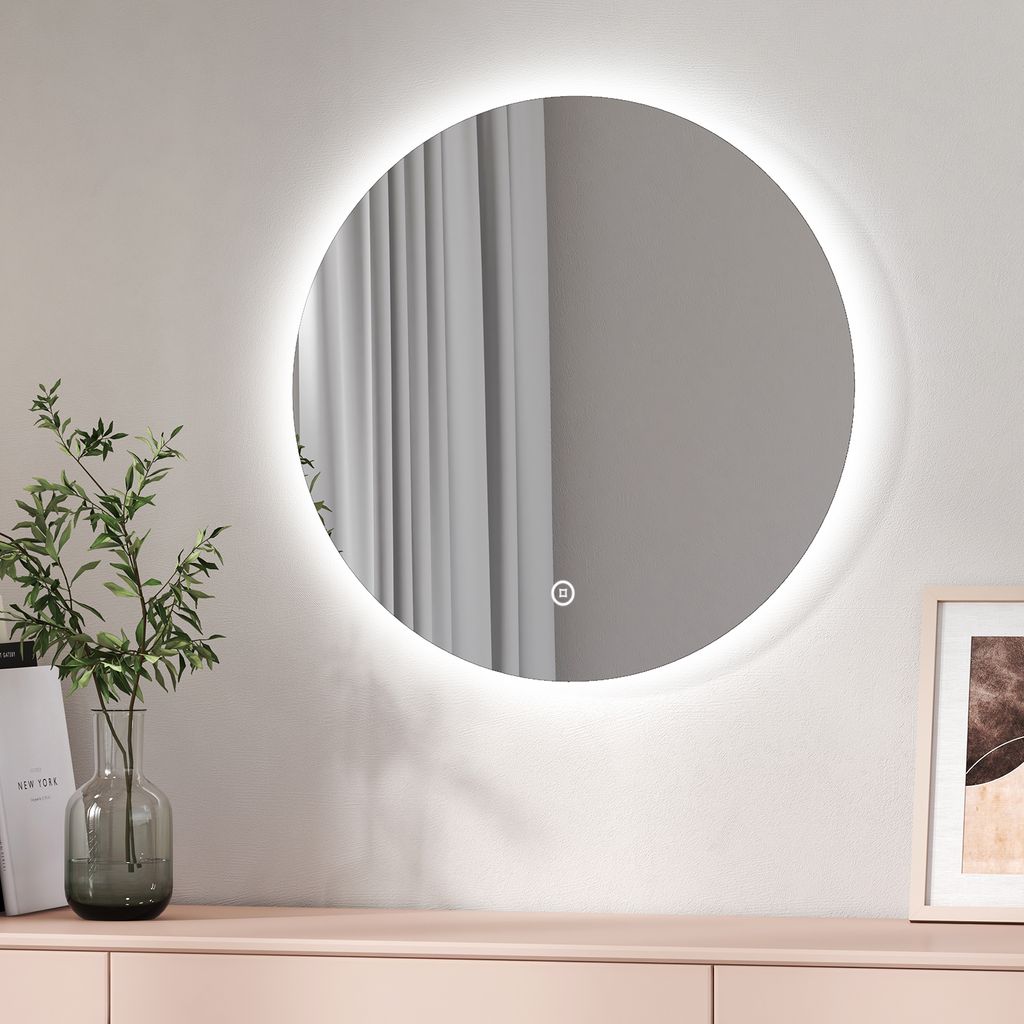A round LED mirror with a touch switch is mounted on a wall above a light pink dresser. A vase with green leaves is placed on the dresser to the left of the mirror. A framed picture is on the right.
