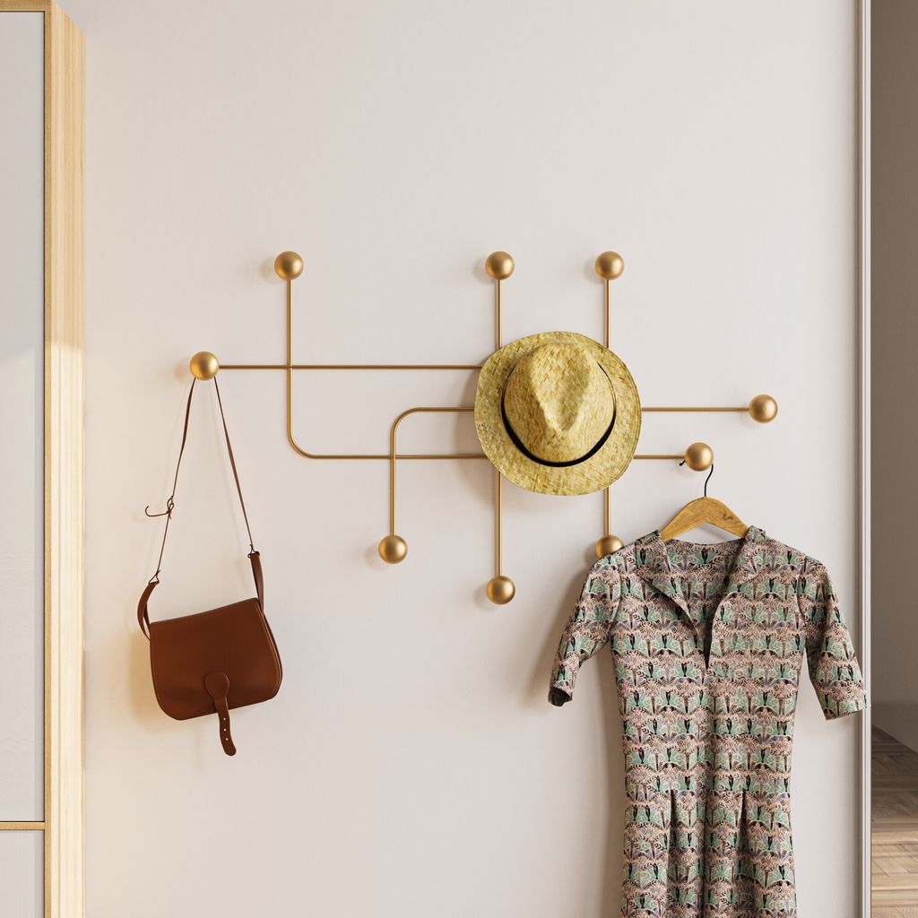 A gold coat rack mounted on a white wall, holding a straw hat, a brown purse, and a patterned dress on a hanger.