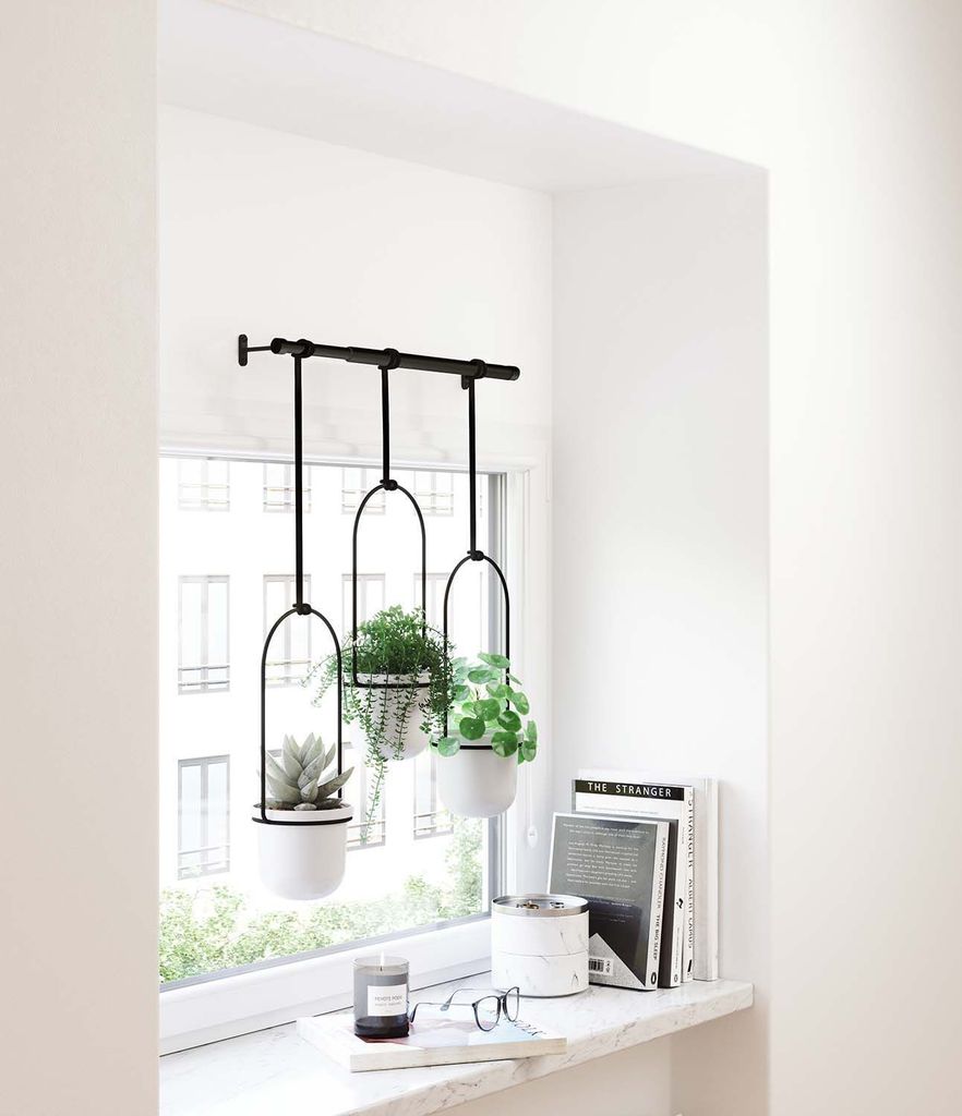 Three hanging planters with plants are displayed on a windowsill next to books and a candle.
