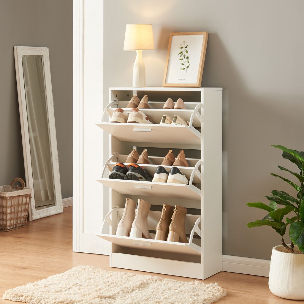 A white shoe cabinet with multiple compartments filled with shoes, placed in a hallway next to a mirror and a plant.
