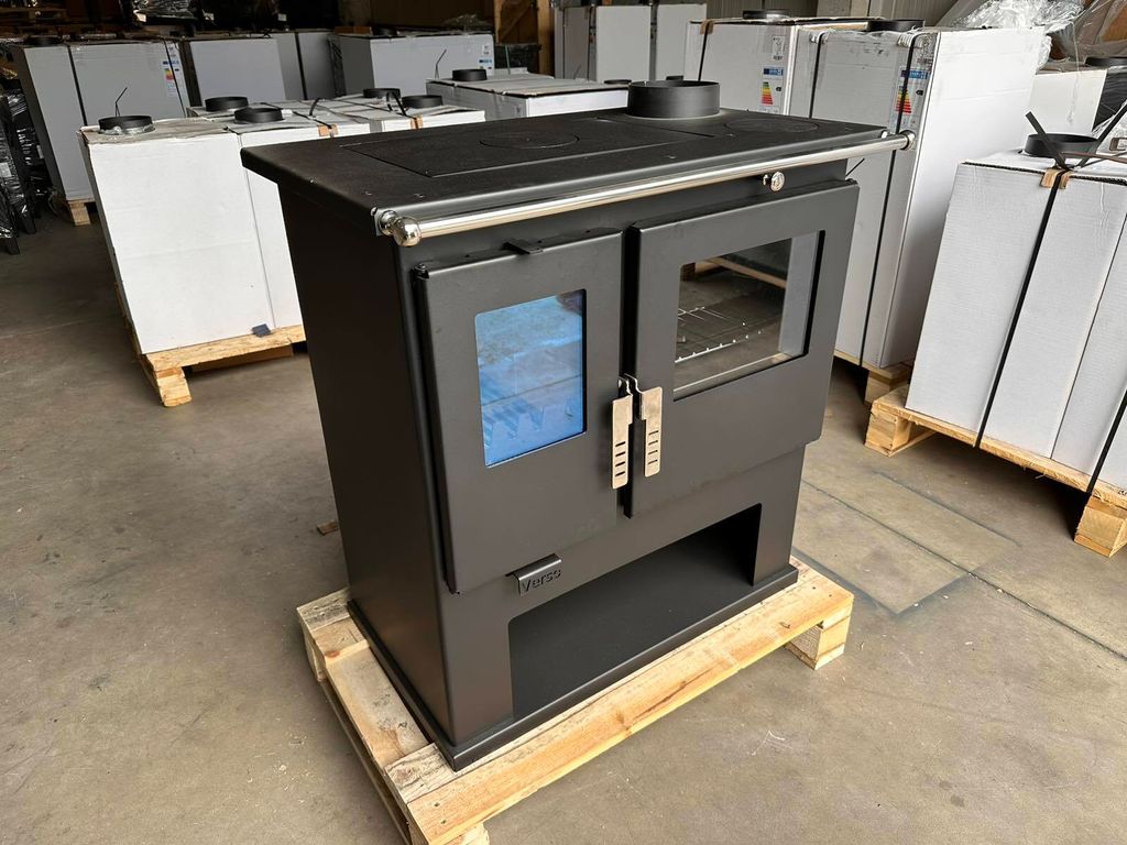 A dark gray wood-burning stove is displayed in a warehouse setting, with other similar stoves or appliances stacked in the background.