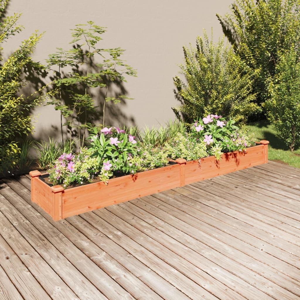 A wooden planter box filled with plants and flowers sits on a wooden deck, with a wall and bushes in the background.