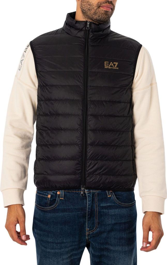 A man wearing a black puffer vest over a beige long-sleeved shirt and blue jeans.