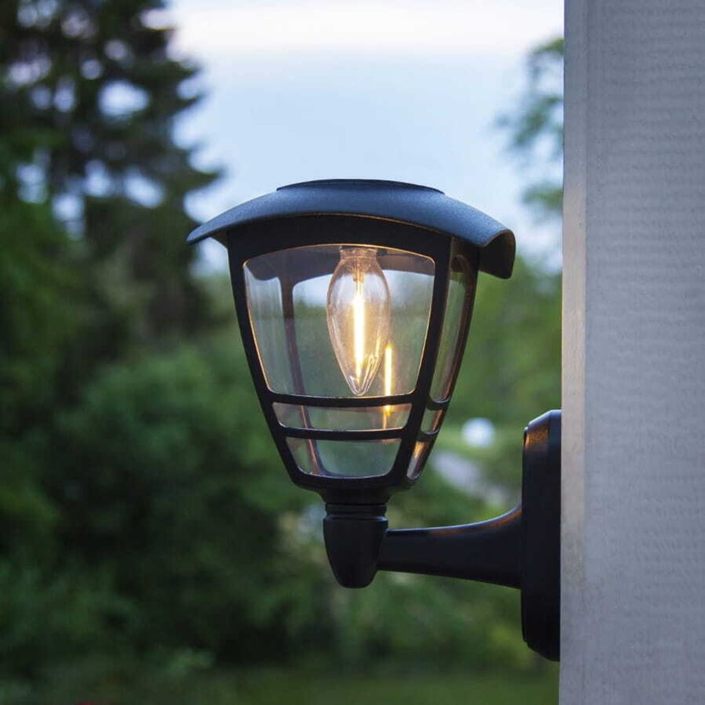 A black outdoor wall lamp with a clear glass shade is mounted on a gray post, with blurred green trees and a light blue sky in the background.