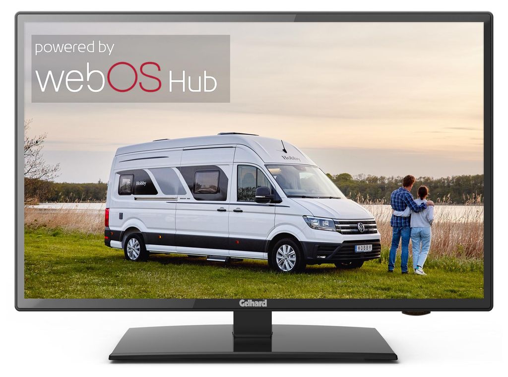 A white camper van parked in a grassy field with a couple standing in front of it, looking out at a body of water. The sky is a soft orange and pink, suggesting sunset or sunrise. The image is displayed on a TV screen.