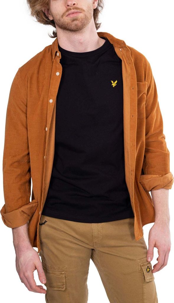 A man wearing a black t-shirt and a brown overshirt, posing against a white background.