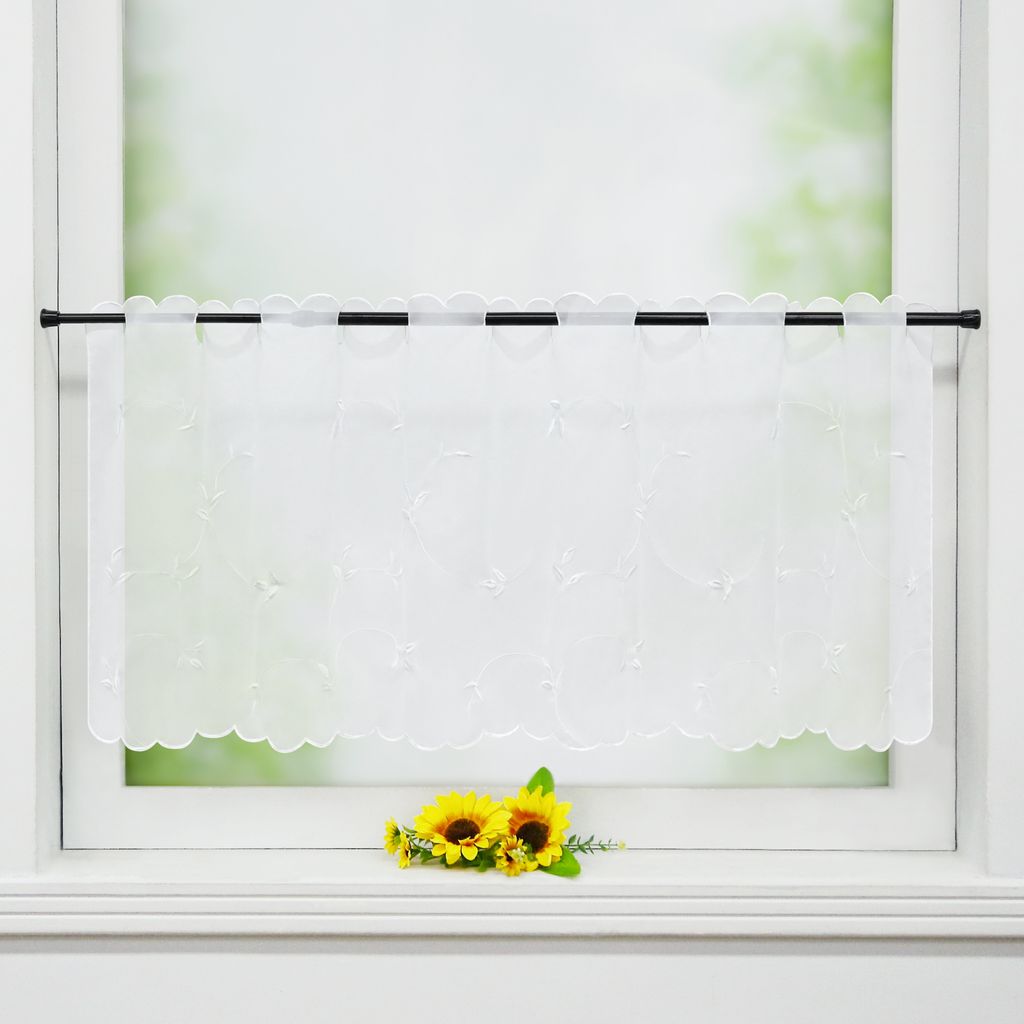 A white sheer curtain with embroidered leaf patterns hangs on a window rod, with two artificial sunflowers placed on the windowsill below.
