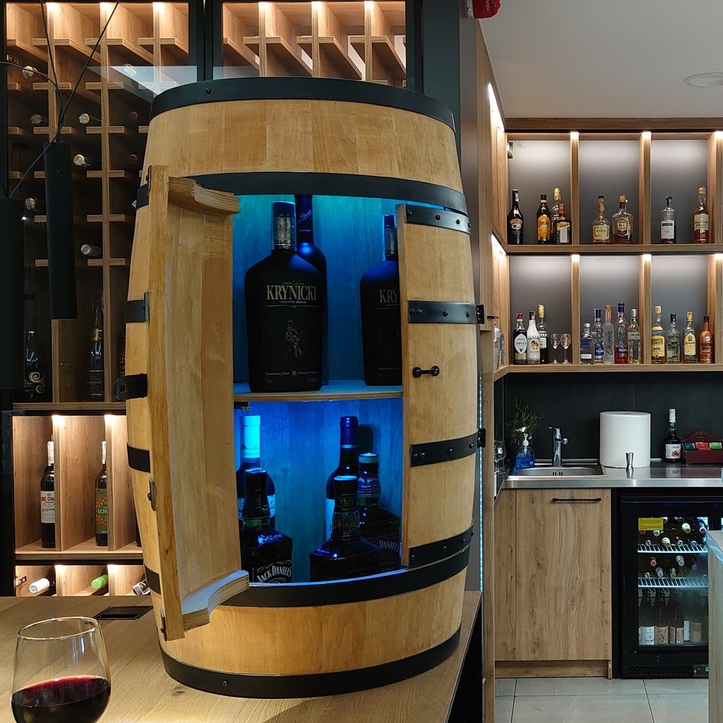 A wooden barrel-shaped bar with bottles of alcohol displayed inside, illuminated by blue LED lights. Shelves with more bottles are visible in the background.