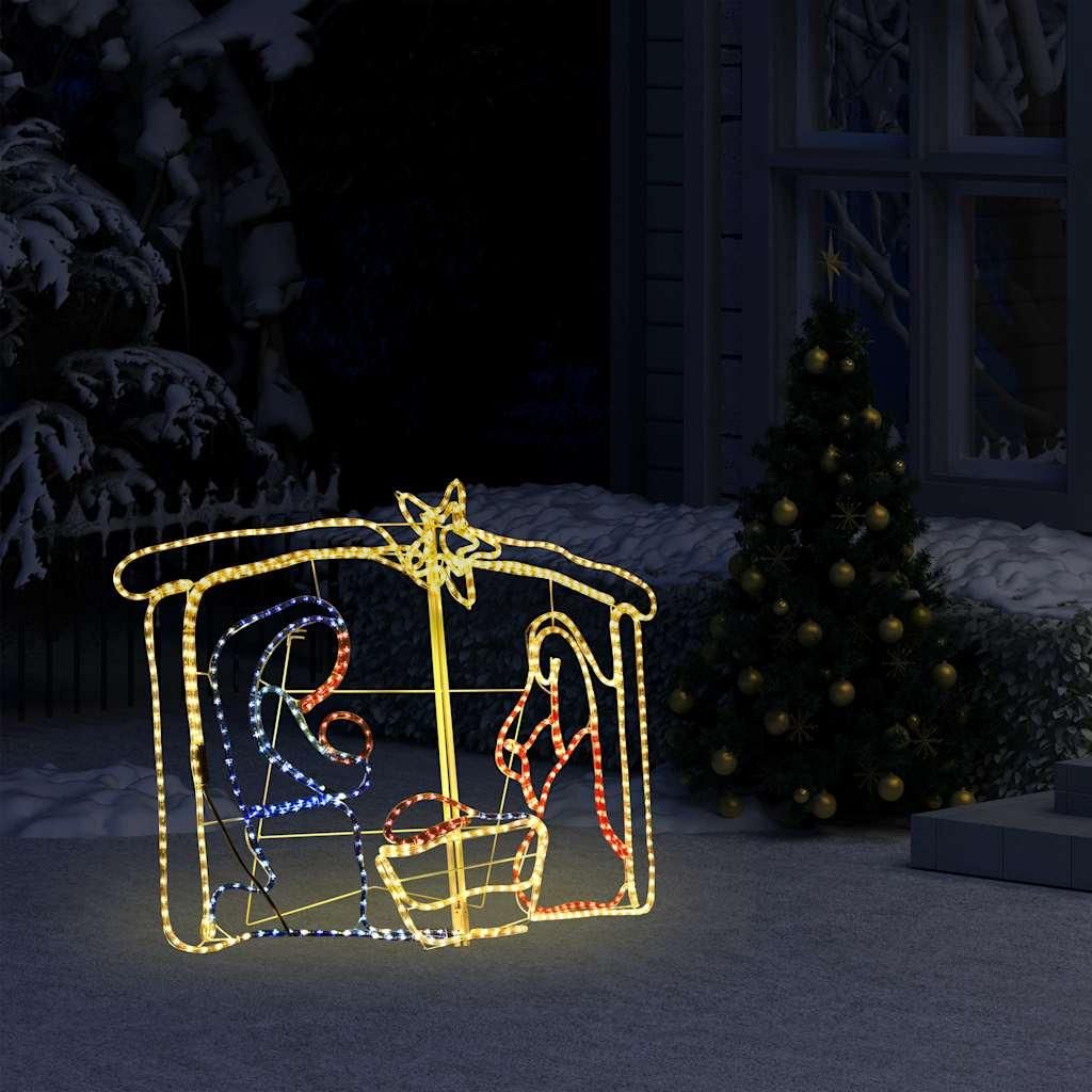A lighted nativity scene decoration is displayed outdoors at night, with a Christmas tree in the background.