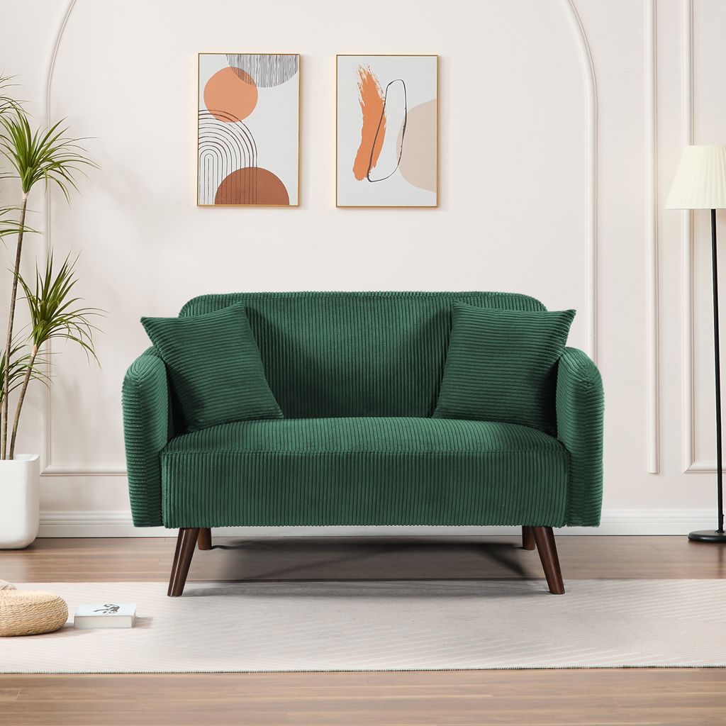 A green corduroy sofa with two throw pillows is shown in a well-lit room with abstract art on the wall and a floor lamp.