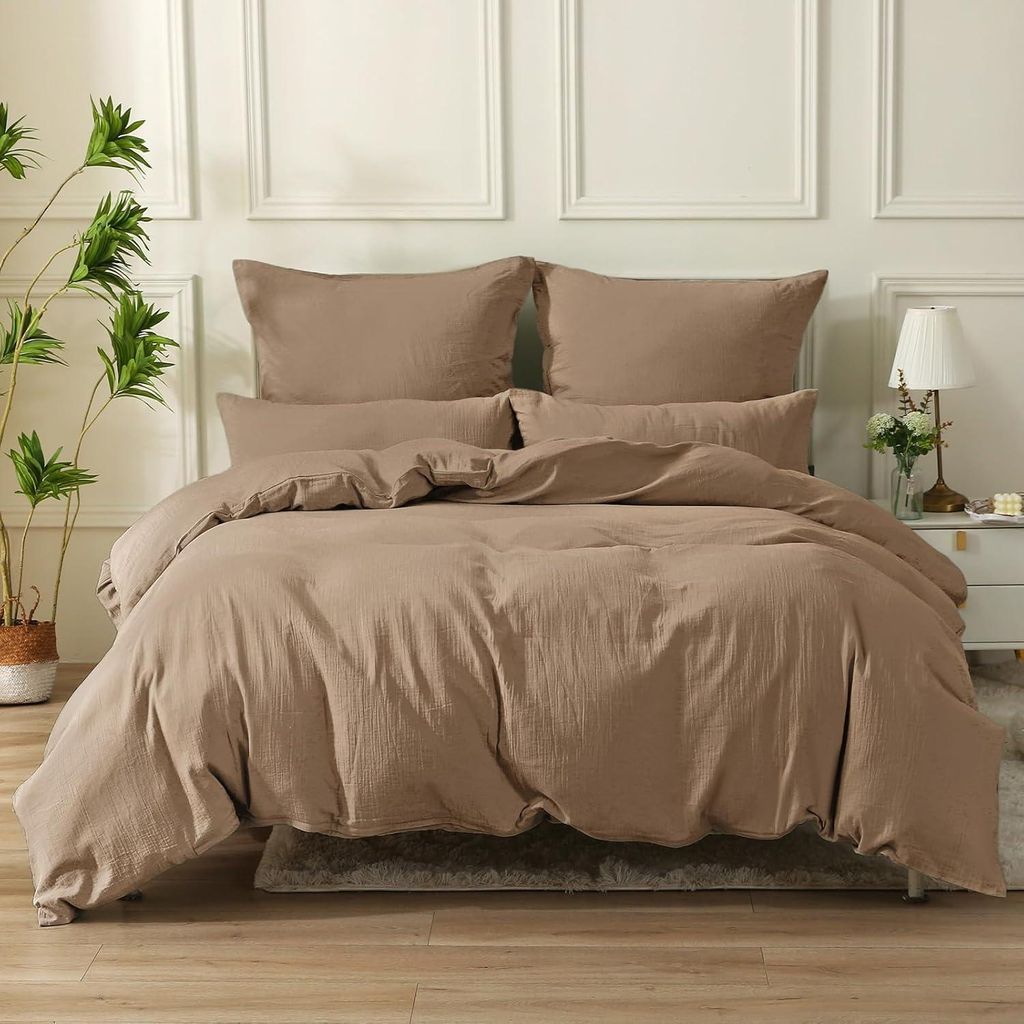 A neatly made bed with a duvet and pillows, set against a wall with decorative molding. A potted plant is visible to the left, and a bedside table with a lamp is to the right.