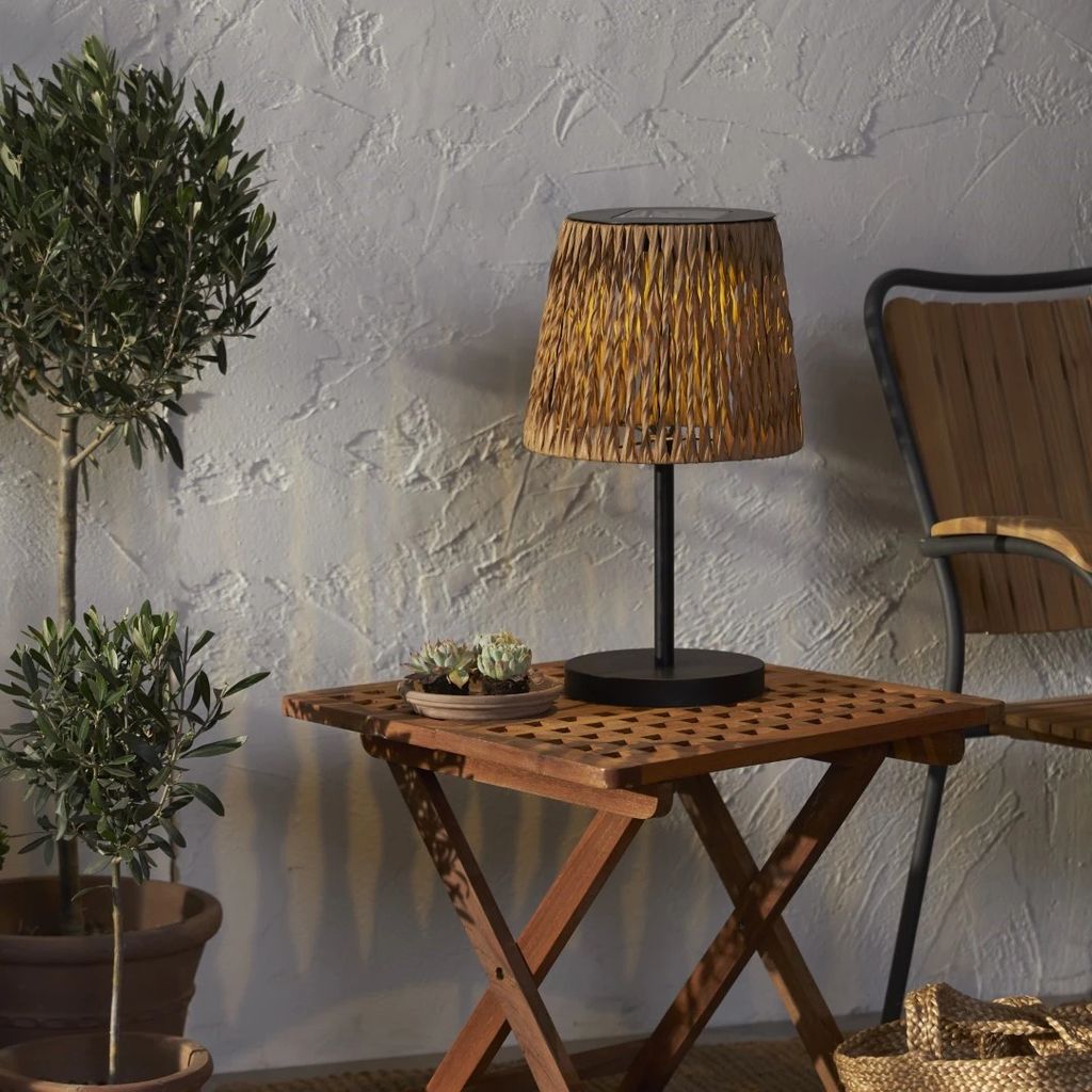 A solar table lamp with a woven shade sits on a wooden table next to potted plants, with a chair in the background.
