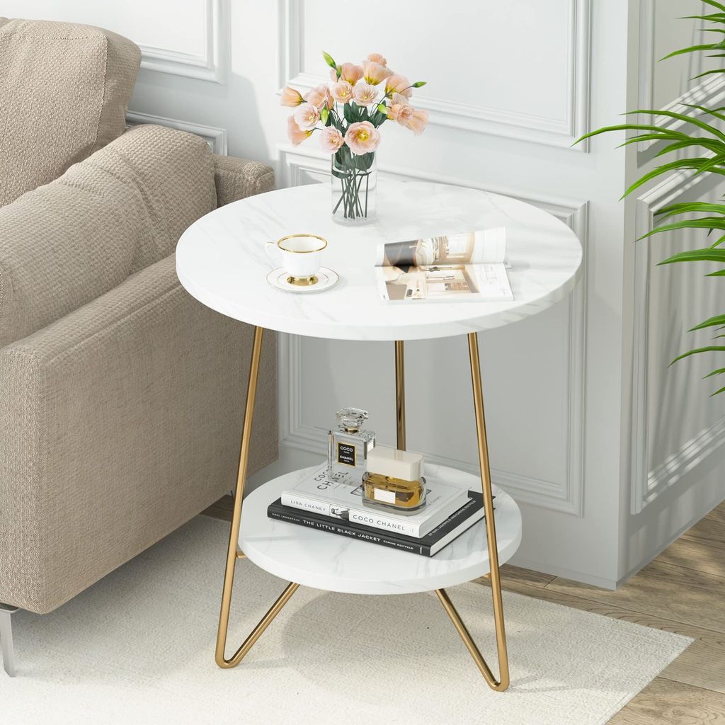 A round, two-tiered side table with a marble-like finish and gold legs, placed next to a sofa in a living room setting. It is decorated with a vase of flowers, a cup and saucer, a book, and perfume bottles.