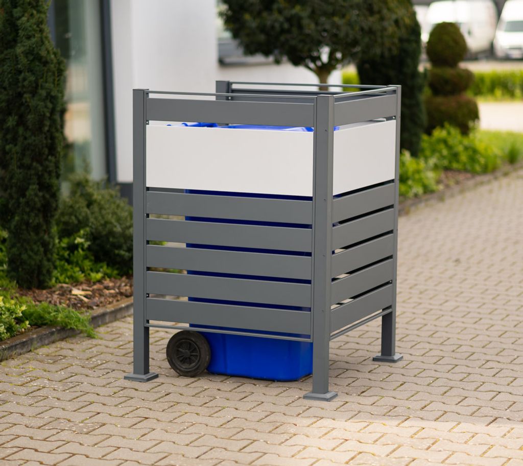 A metal trash can enclosure with a blue bin inside, placed on a paved walkway next to greenery.