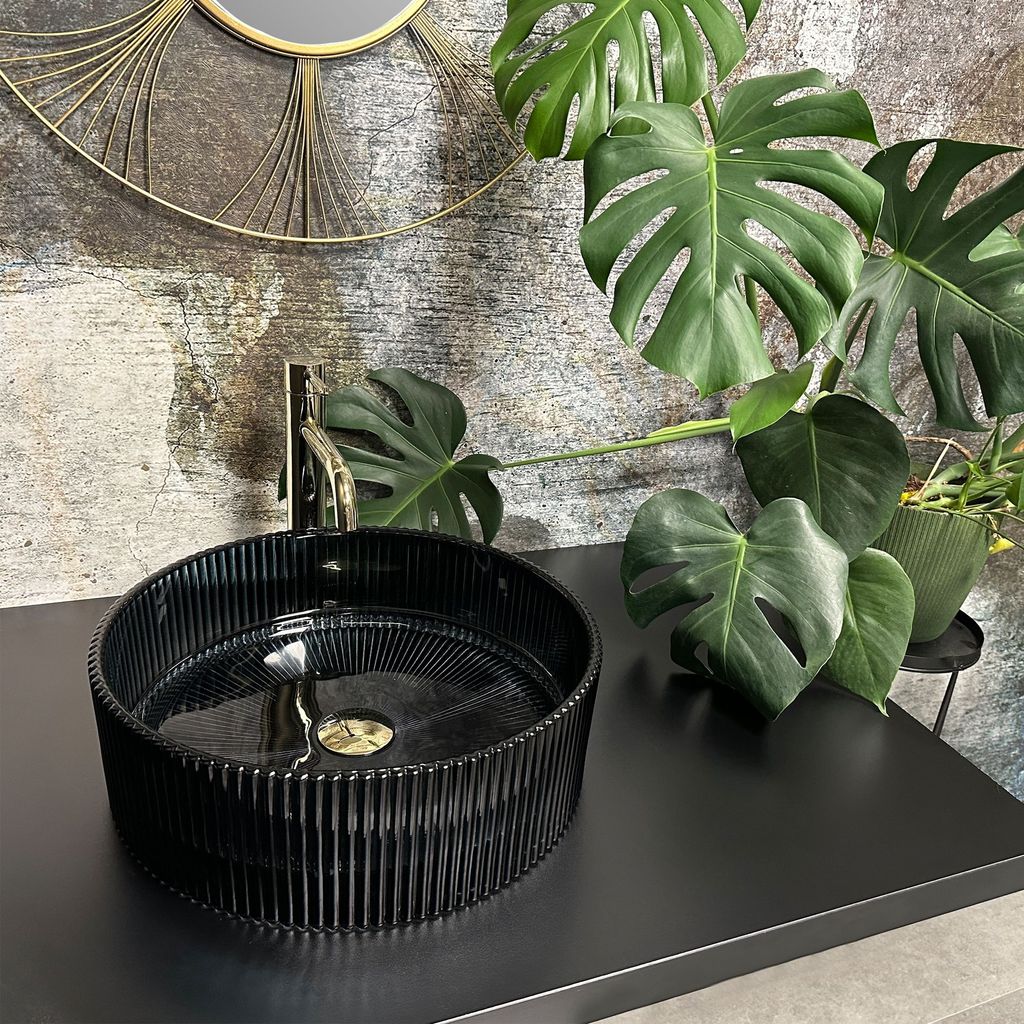 A black basin with a gold faucet sits on a black countertop, with a large Monstera plant and a decorative mirror in the background.