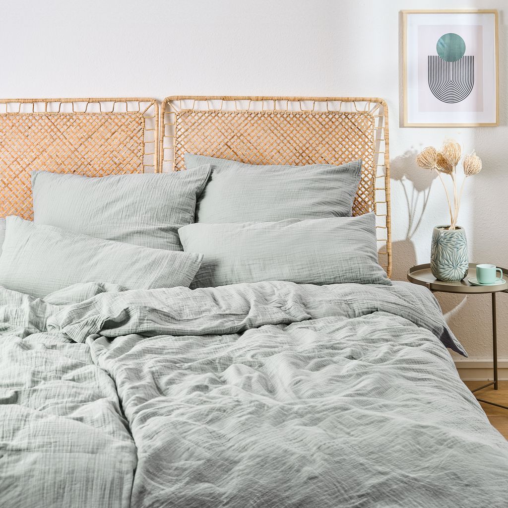 A neatly made bed with light blue musselin bedding and a woven headboard.