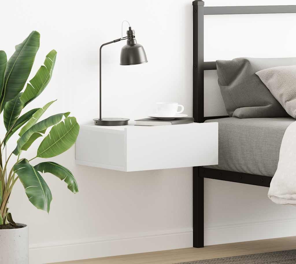 A white floating nightstand is mounted on a white wall next to a bed. A black desk lamp and a white teacup are on top of the nightstand. A large potted plant is to the left of the nightstand.