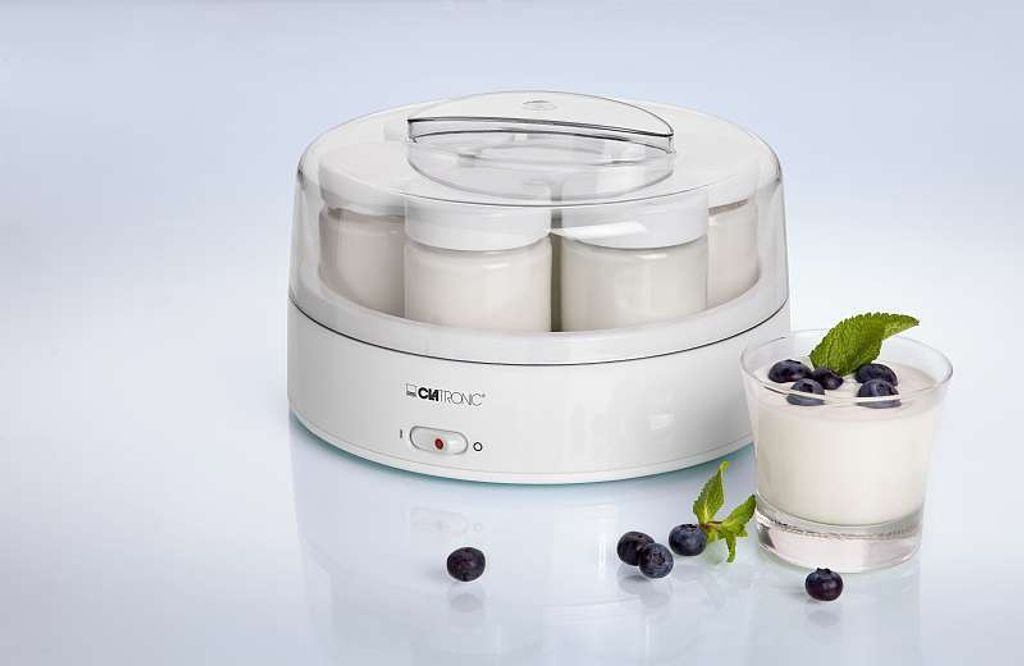 A white yogurt maker with multiple jars filled with yogurt, and a glass of yogurt topped with blueberries and a mint leaf, set against a light gray and white gradient background. Several blueberries are scattered around the foreground.