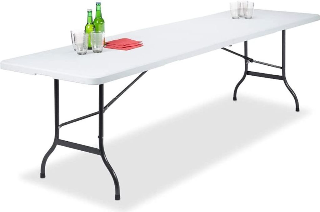 A long, white folding table with black legs, set up on a white background. There are a few glasses, bottles, and red napkins on the table.