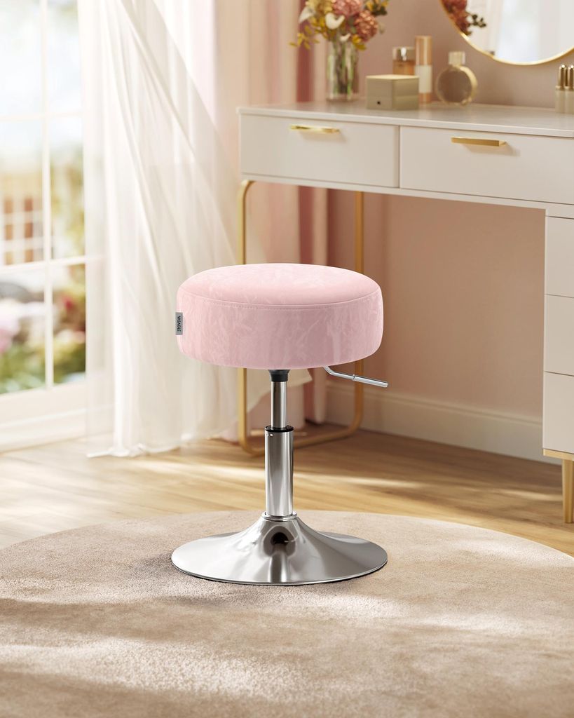 A pink velvet stool with a silver adjustable base is placed on a light beige rug in front of a white vanity desk with gold accents.