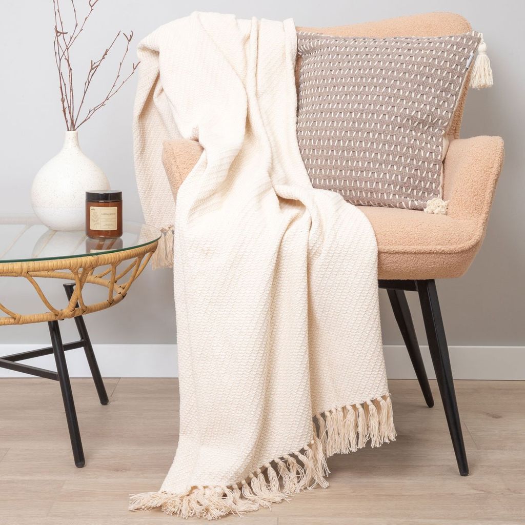 A cream-colored throw blanket is draped over a chair next to a small table with a vase and a candle.