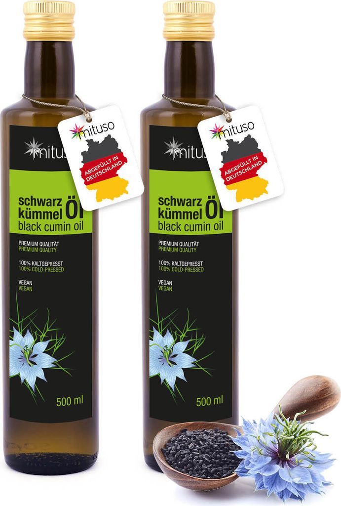 Two bottles of black cumin oil are displayed side-by-side, with a small wooden bowl of black cumin seeds and a blue flower in the foreground.
