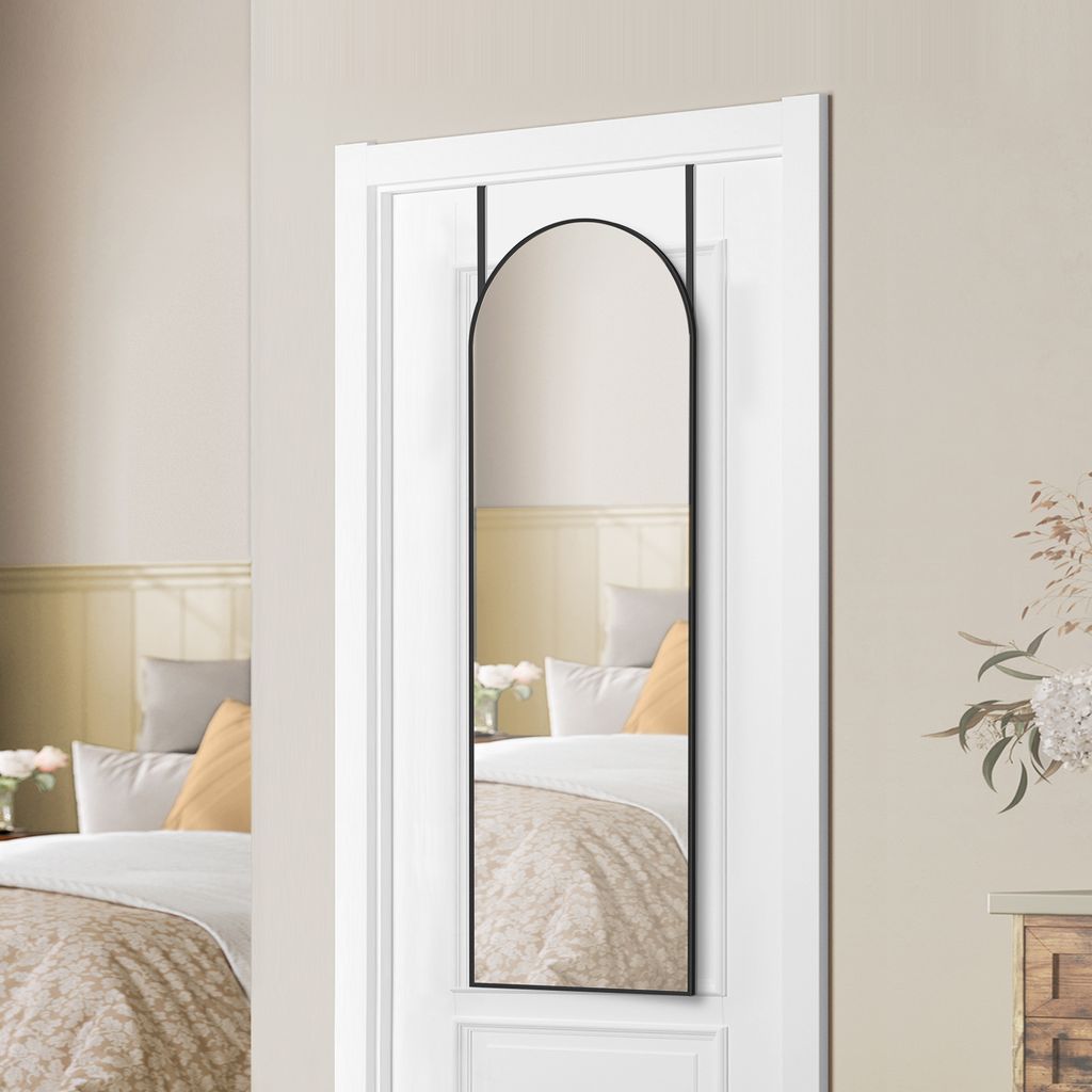 A full-length mirror with a black arched frame hangs on a white door, reflecting a bedroom scene with a bed and pillows.