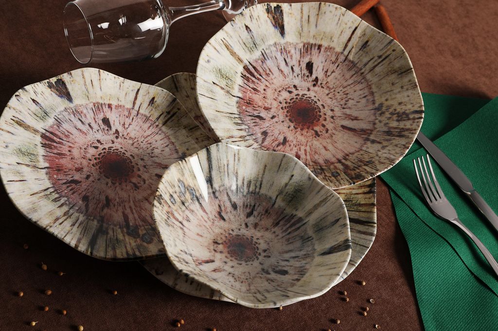 A set of decorative plates and bowls arranged on a brown surface, with a wine glass in the background and cutlery and a green napkin to the side.