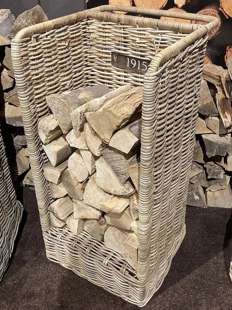 A wicker basket filled with firewood, placed on a brown carpet with stacks of firewood in the background.