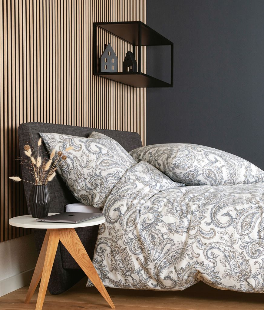 A bed with patterned bedding and pillows, next to a small wooden side table with a vase of dried flowers and a laptop.