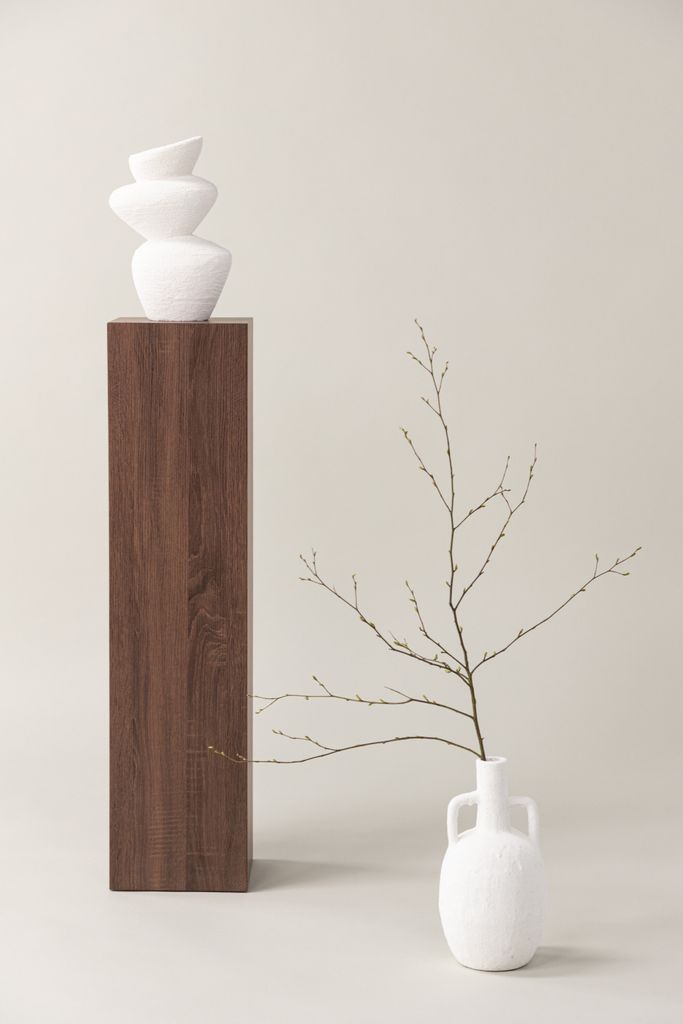 A minimalist studio shot featuring two white vases and a wooden pedestal against a light gray background.