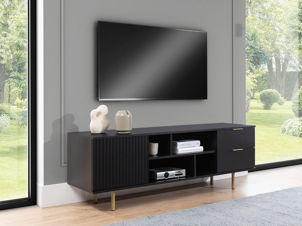 A modern living room with a TV mounted on the wall above a black TV stand with gold legs. The TV stand has a fluted door, drawers, and open shelves. A large window with a view of a garden is on the right.