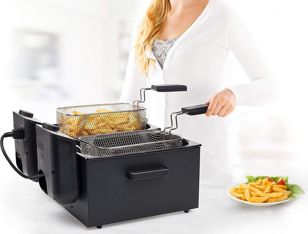 A woman is using a black deep fryer that contains french fries. Another plate of french fries is on the right.