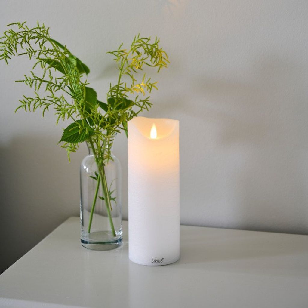 A white LED candle and a vase with green foliage sit on a white surface against a light gray wall.