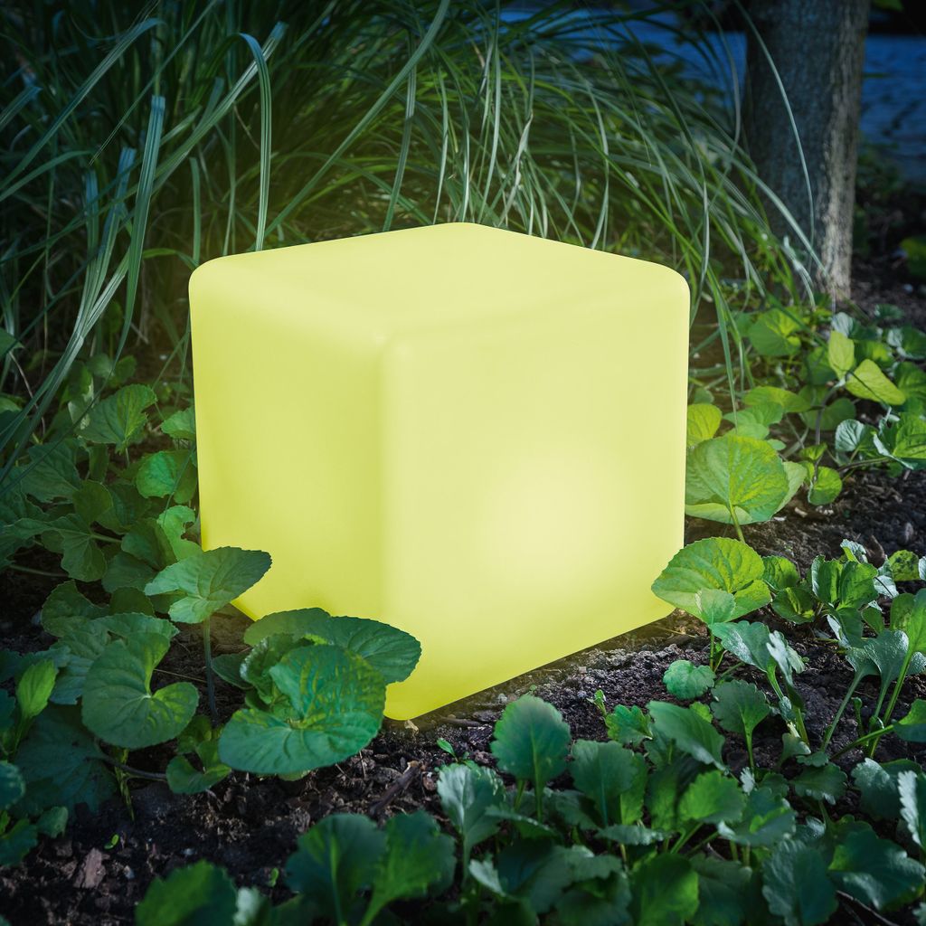 A glowing yellow cube-shaped solar light sits in a garden surrounded by plants and grass.
