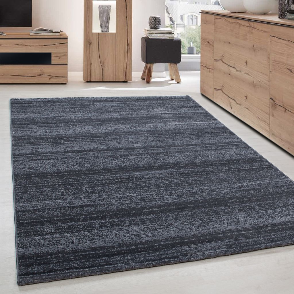 A dark gray rug with a subtle linear texture is laid out on a light-colored floor in a modern living room setting. Furniture pieces, including a wooden cabinet and a small stool, are visible in the background.