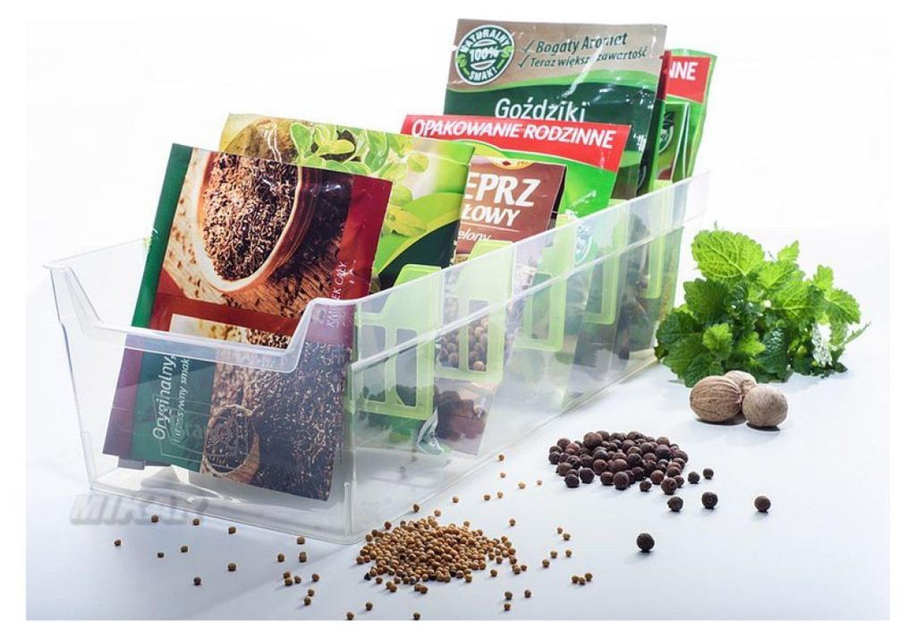 A clear plastic organizer filled with various spice packets, with loose spices and nutmeg scattered around it on a white surface. Fresh mint leaves are also visible.