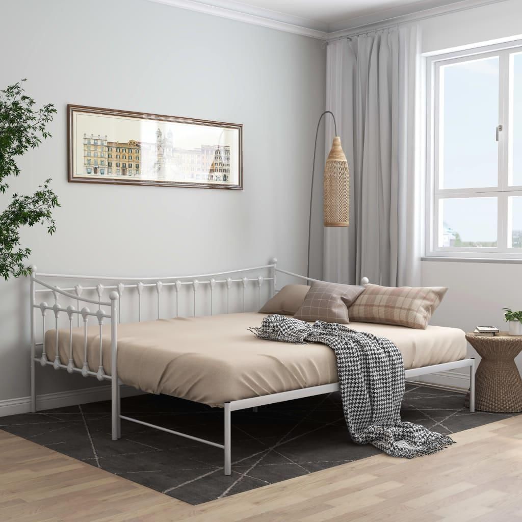 A white metal daybed with a beige mattress and pillows is shown in a room with a large window and curtains.