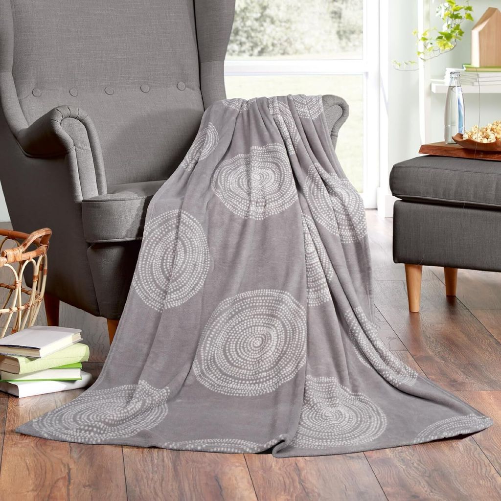 A gray blanket with a circular pattern is draped over a gray armchair in a living room setting. Books are stacked on the floor next to the chair, and a small ottoman with a tray of snacks is visible in the background.