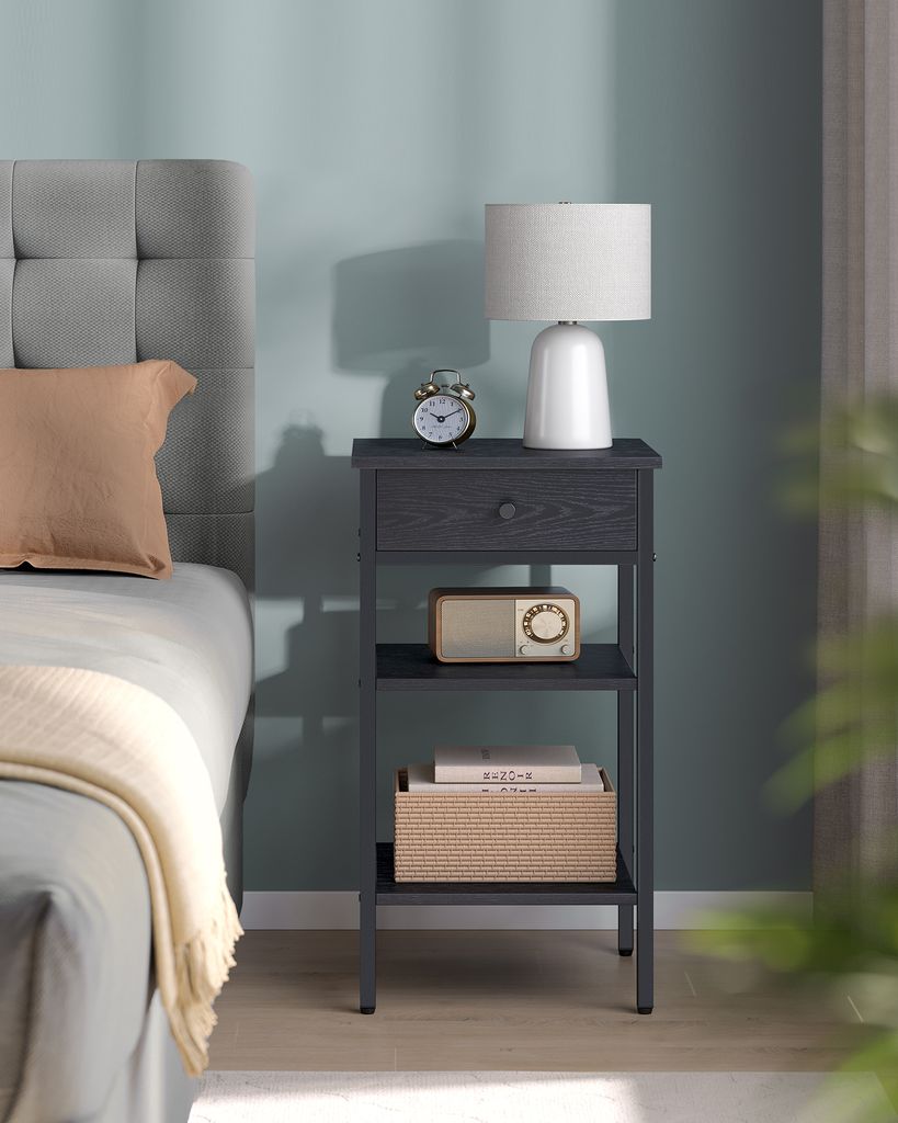 A bedside table with a lamp, alarm clock, radio, and books next to a bed.