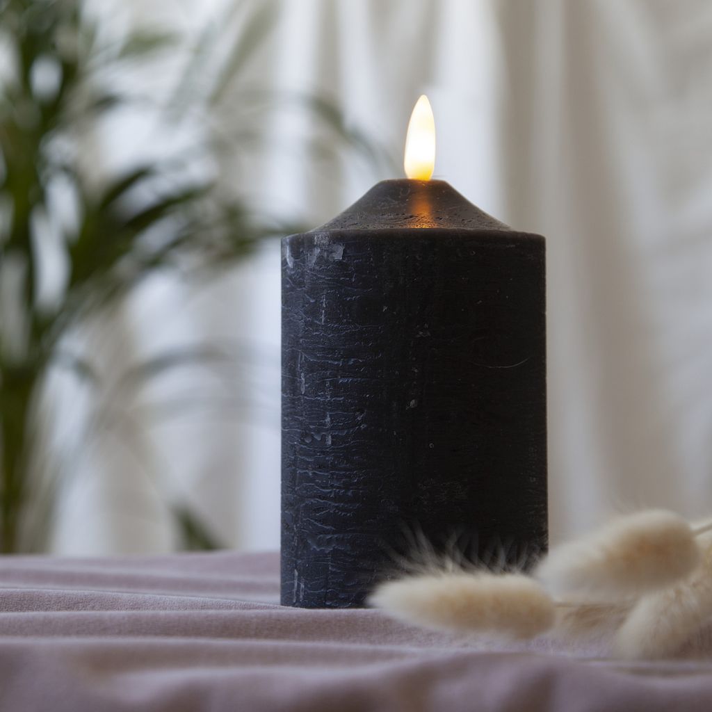 A black LED candle with a flickering flame sits on a textured surface, with a blurred plant and curtain in the background.