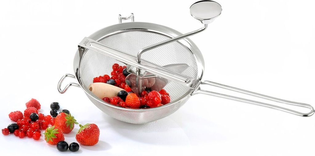 A stainless steel food mill filled with berries, with some berries scattered around it on a white background.
