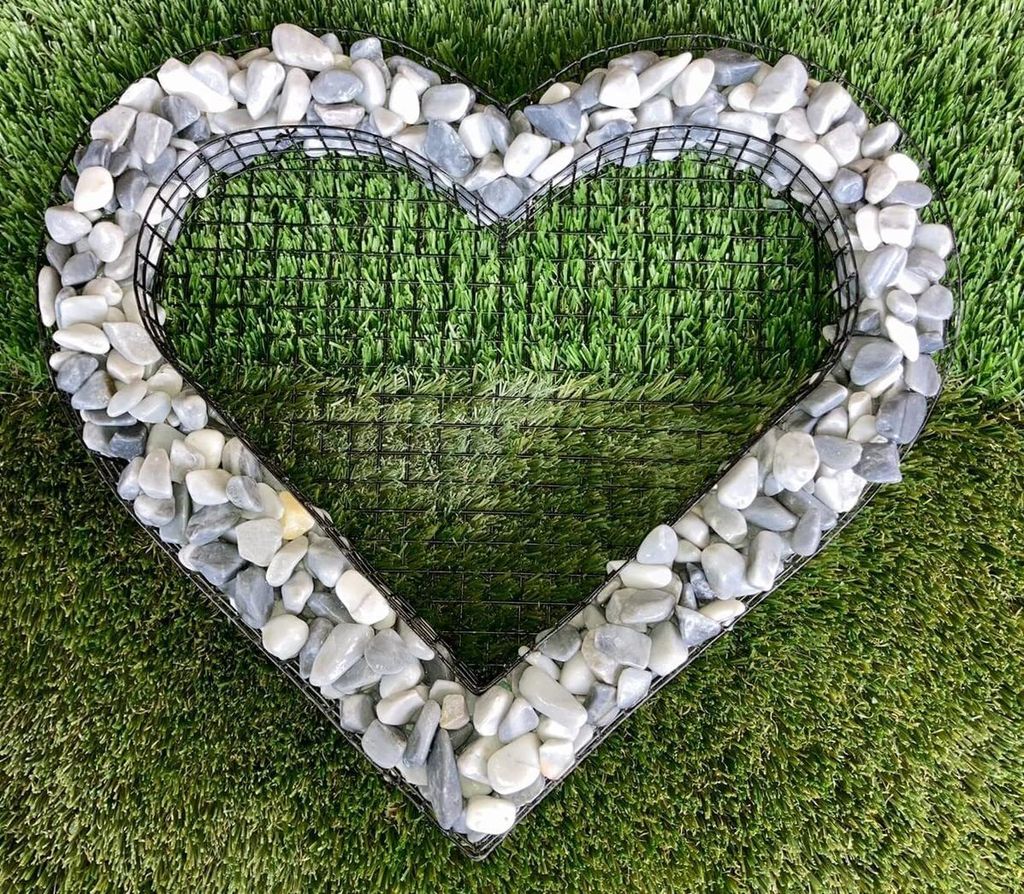 A heart-shaped wire frame filled with grey and white stones, placed on artificial grass.