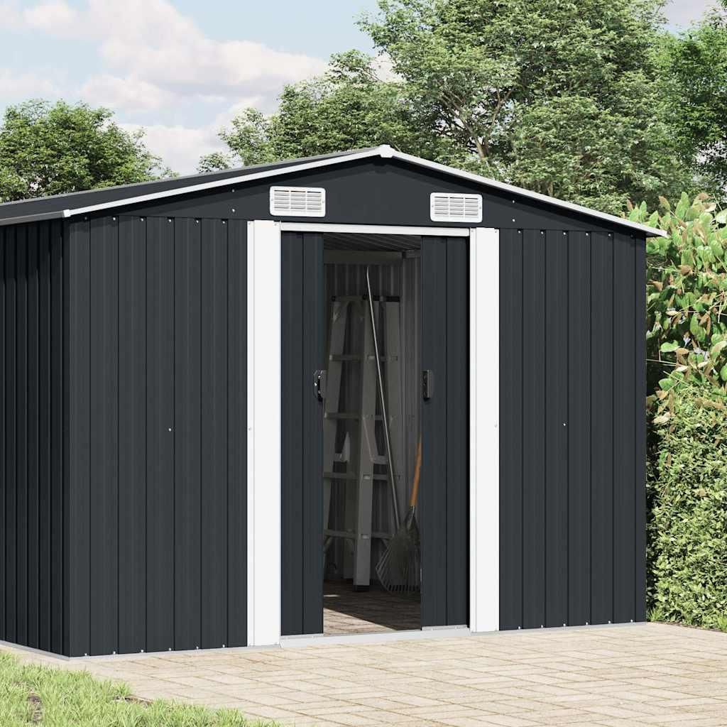 A dark gray metal shed with white trim and open doors, set against a backdrop of green trees and a cloudy sky. The shed is situated on a paved patio.