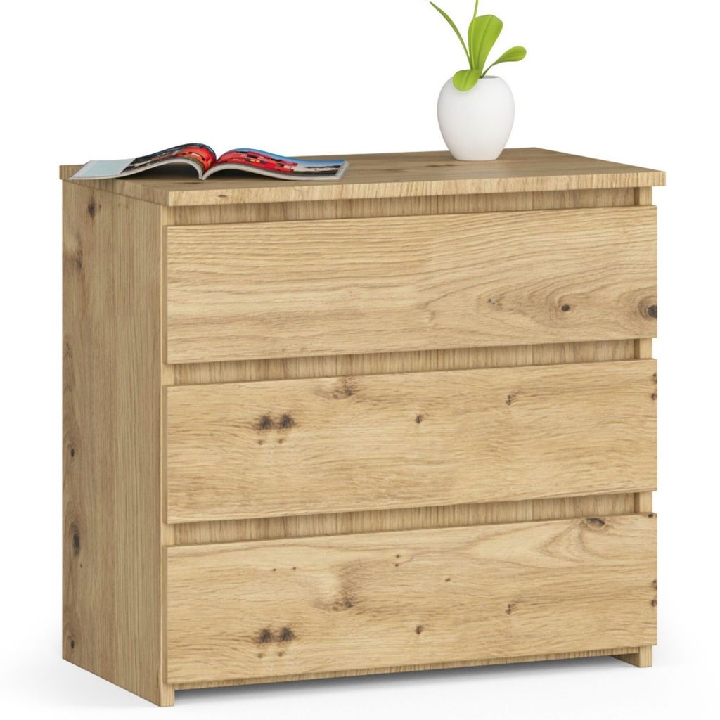 A wooden chest of drawers with three drawers, a magazine and a small plant in a white vase on top, against a white background.